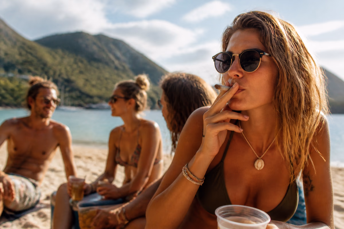 Mujer con gafas de sol fumando un porro en una playa tropical con amigos con bebidas al fondo