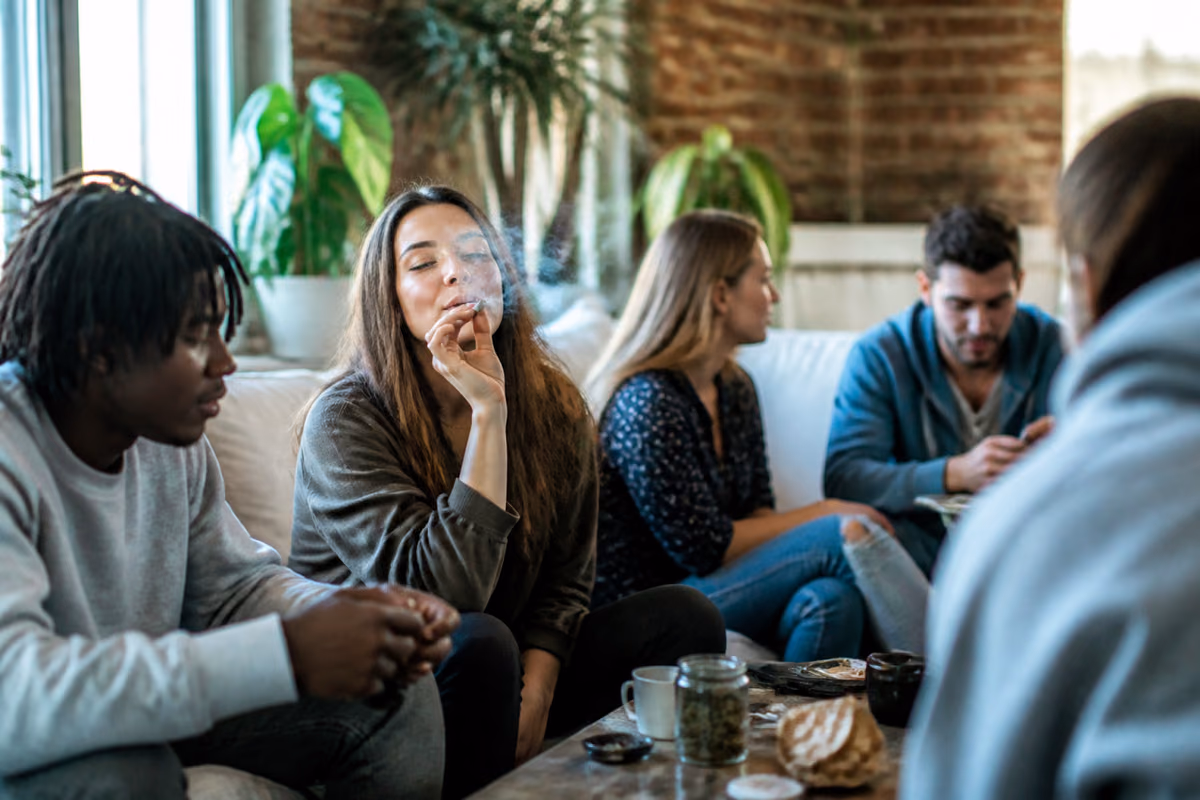 Jóvenes adultos en un sofá mientras una mujer fuma un porro, con un frasco de cogollos de cannabis en la mesa