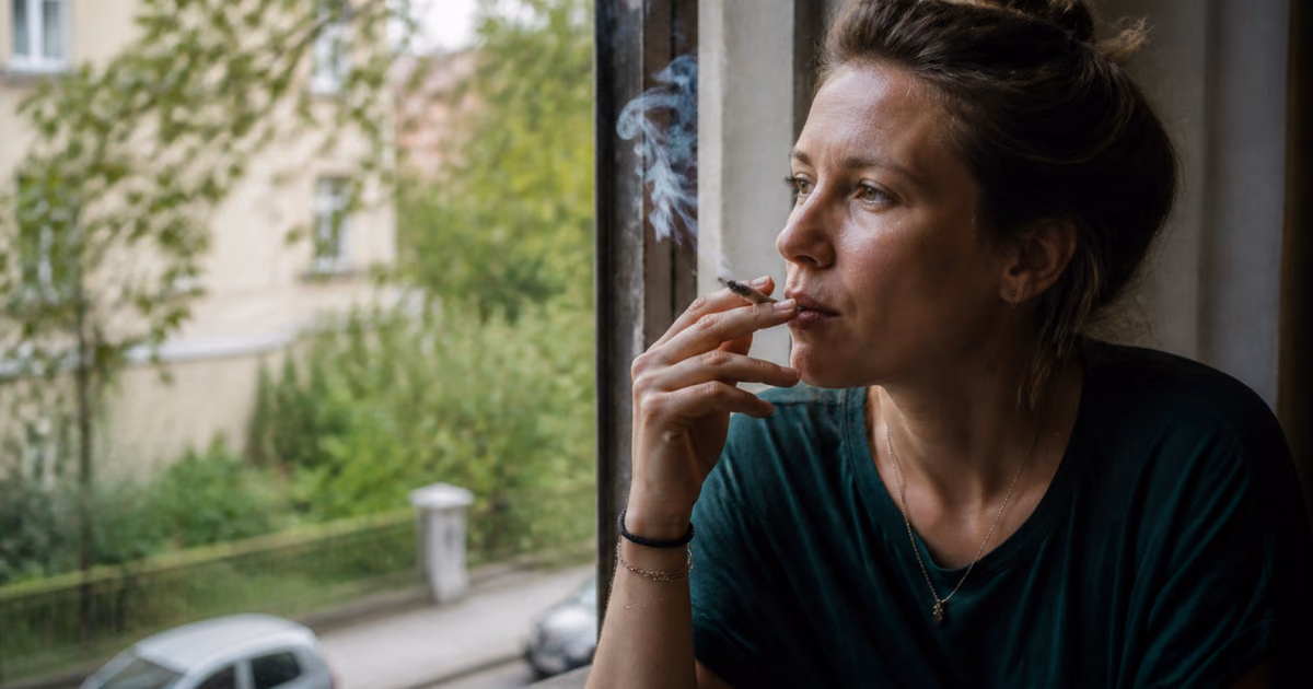 Mujer fumando un porro junto a una ventana y mirando una calle arbolada